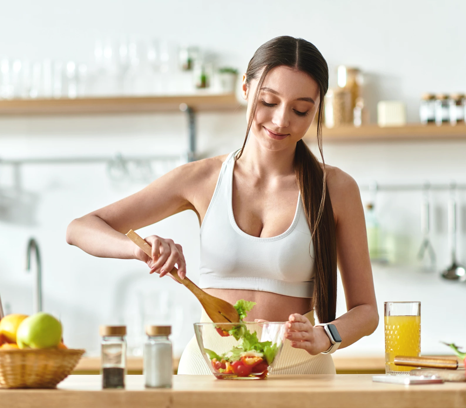 Woman in a sports bra tossing healthy chicken salad with cherry tomato and lettuce, with a glass of orange juice on the counter.