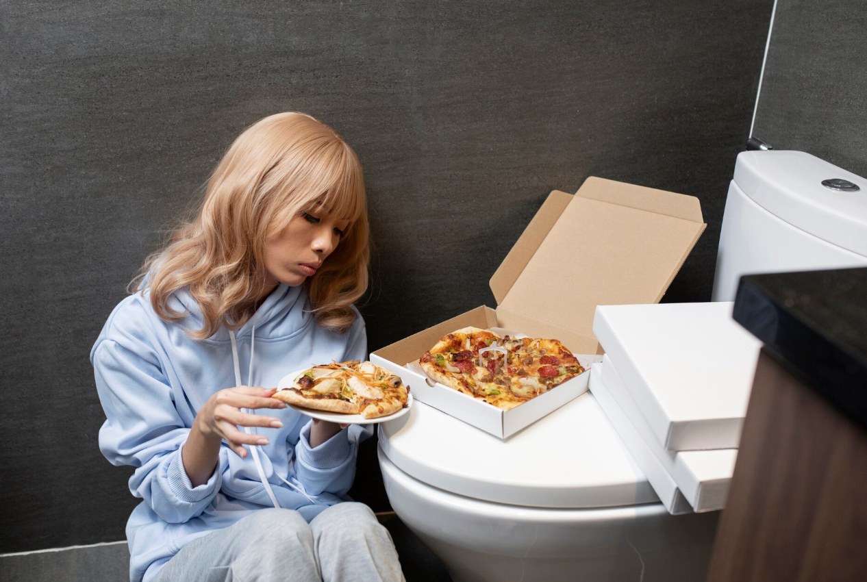 A person sits on a bathroom floor near pizza boxes, binge-and-purge behavior of bulimia nervosa.