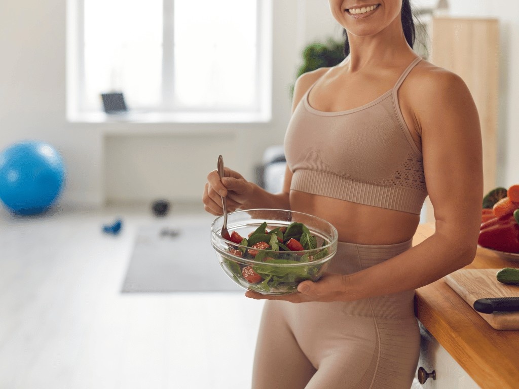 Woman wearing beige sports bra and leggings stands in kitchen holding a glass bowl of salad with tomatoes and greens.