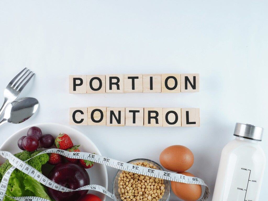 Bowl of fruits, eggs, and water bottle beside measuring tape with wooden blocks spelling “portion control” for healthy eating.