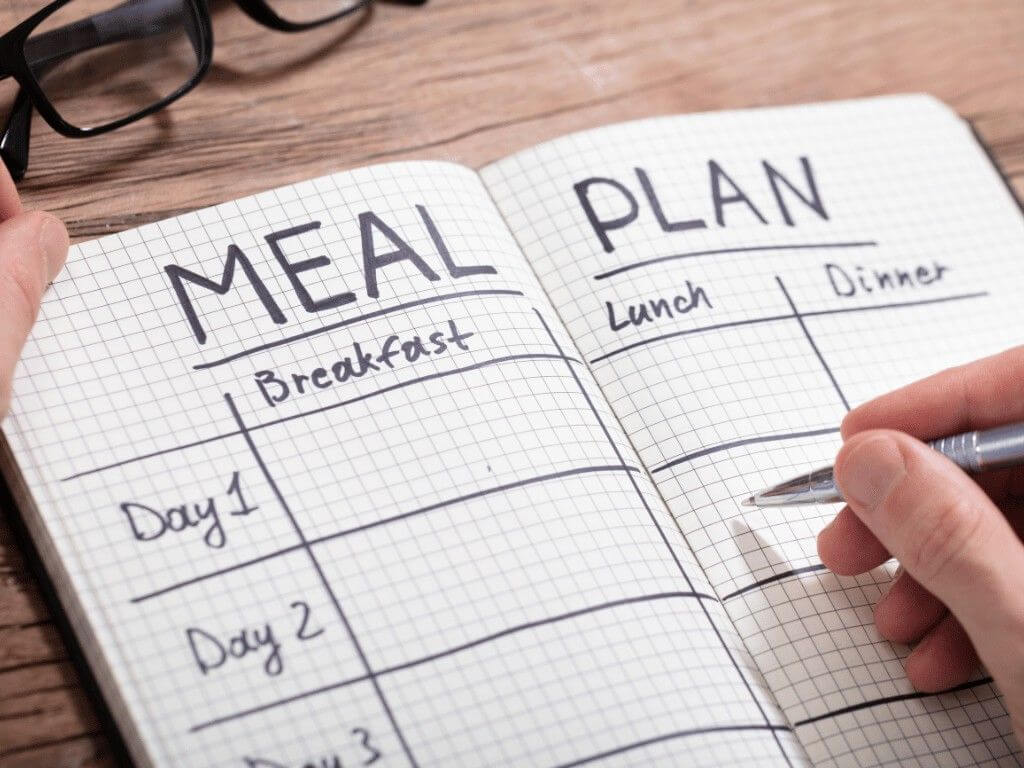 Hand writing in a meal planning notebook to create a weekly gluten free meal schedule.