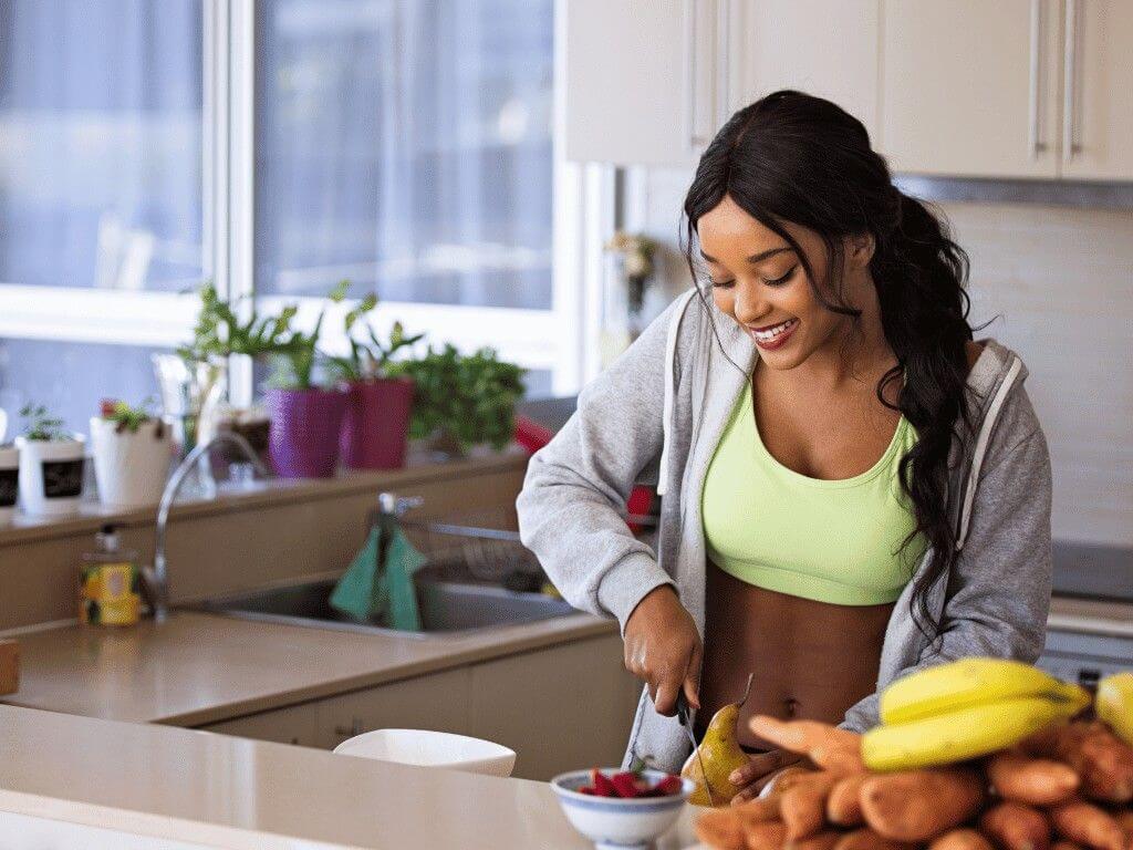 Woman in activewear cuts fruit in kitchen as part of building consistency and healthy habits in 75 Soft Challenge.
