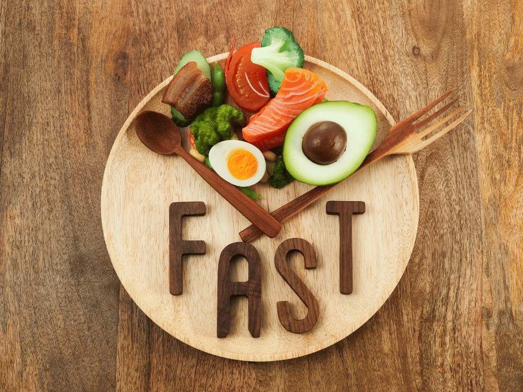 plate with words 'fast', spoon and fork representing clock hands and some foods in a corner. It serves as an indication of the intermittent fasting practice