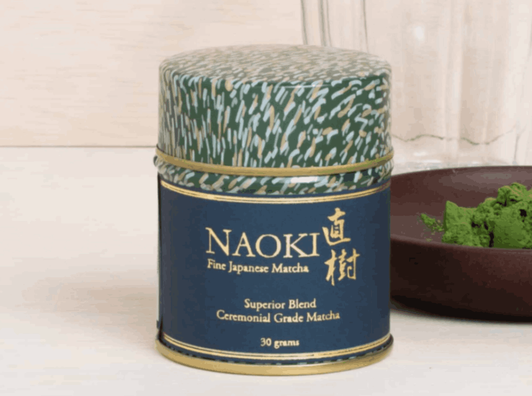 Naoki Matcha tin with bowl of organic green tea powder and glass in background.
