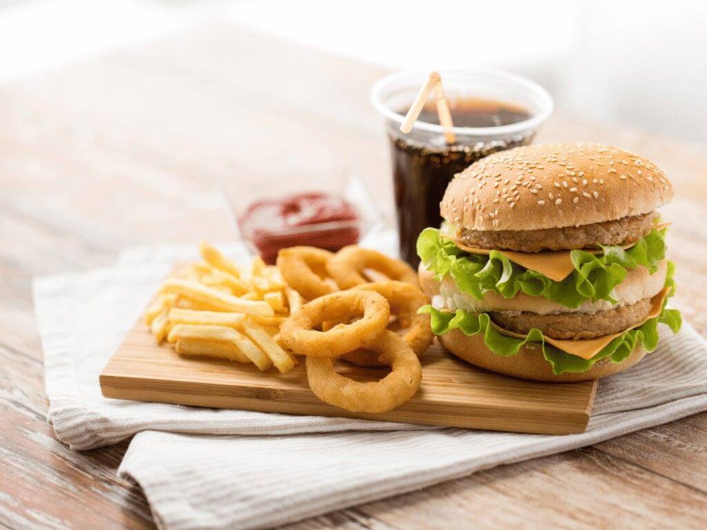 Burger, fries, onion rings, and soda on a wooden board showing processed fast food to limit for a healthier diet