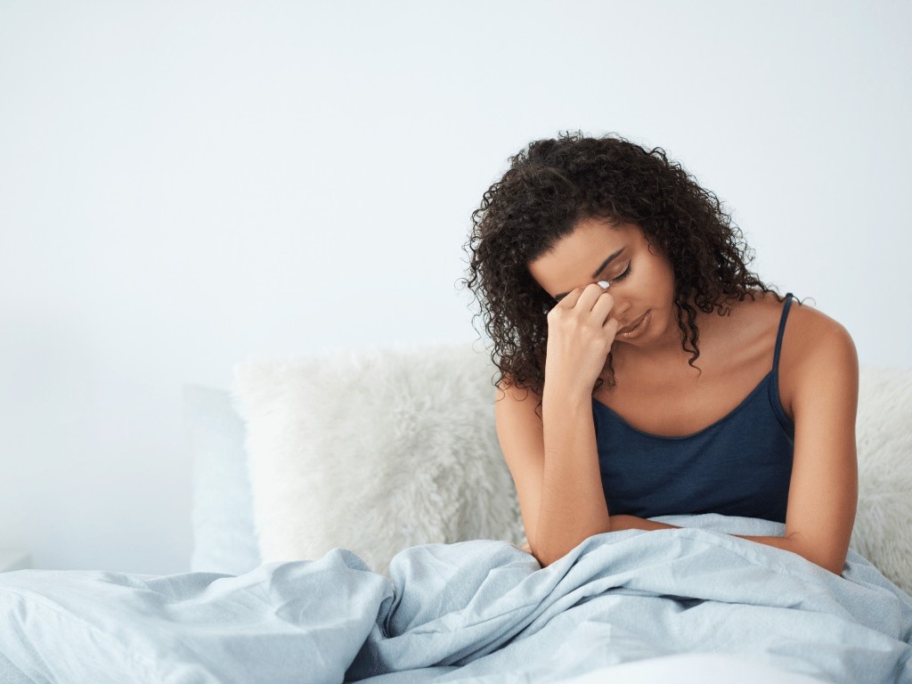 Tired woman sitting on bed experiencing fatigue and headaches as OMAD diet side effects.