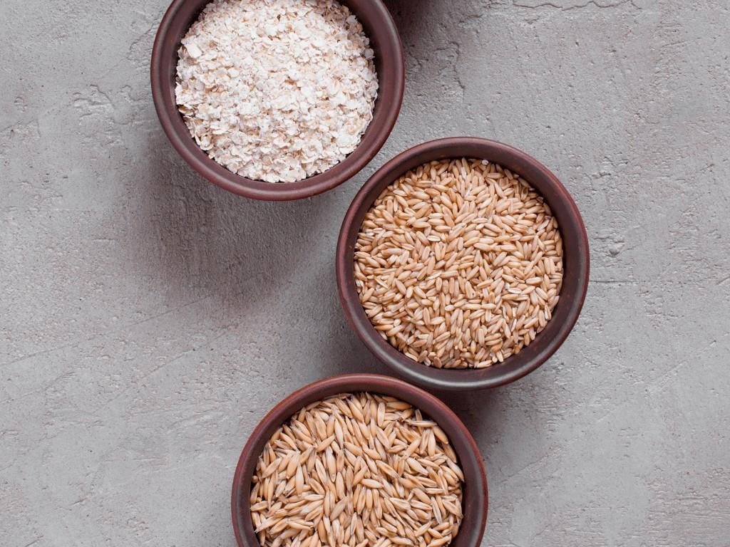 Three bowls with oat flour, rolled oats, and whole oat groats on gray surface, showing oatmeal types for healthy weight loss