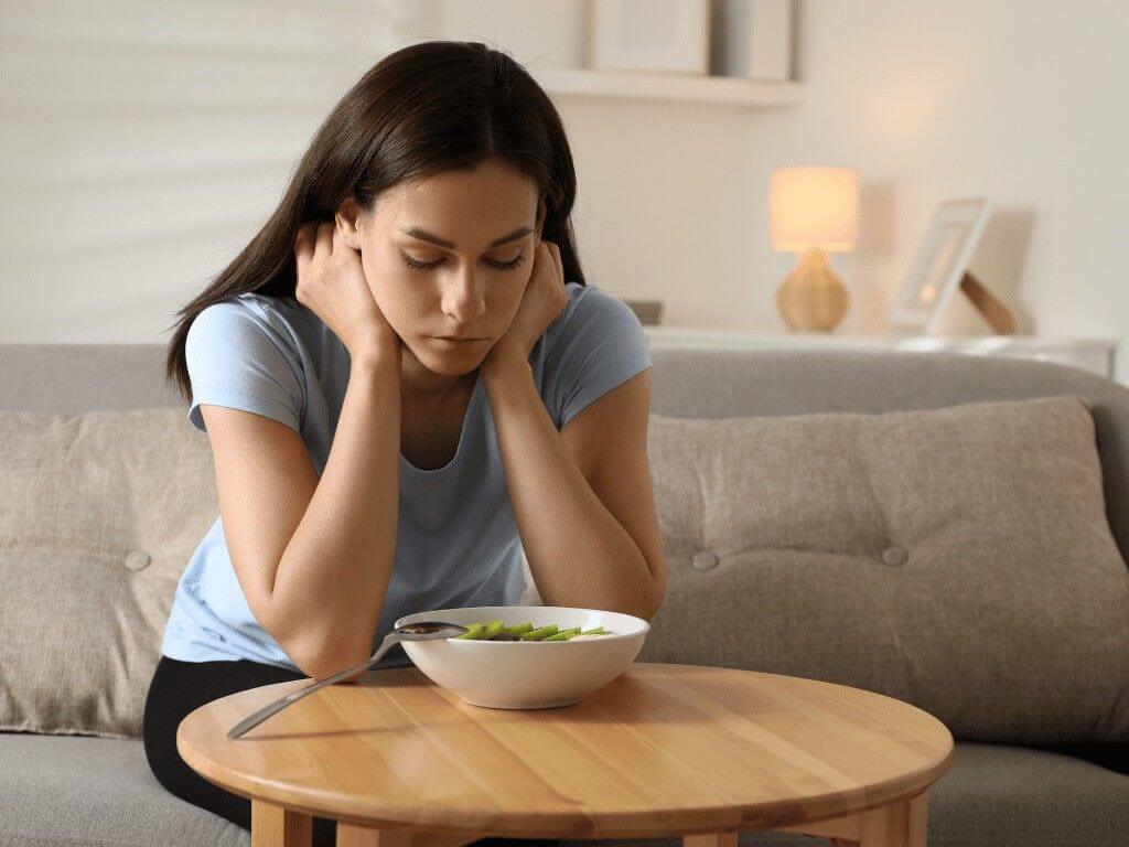 Woman sitting on couch looking down at a bowl of salad, showing signs of distress linked to eating disorder awareness.