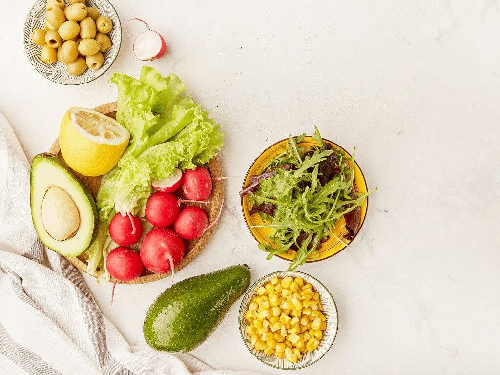 Fresh low FODMAP diet ingredients including avocado, lettuce, radish, corn, lemon, and olives on a table.
