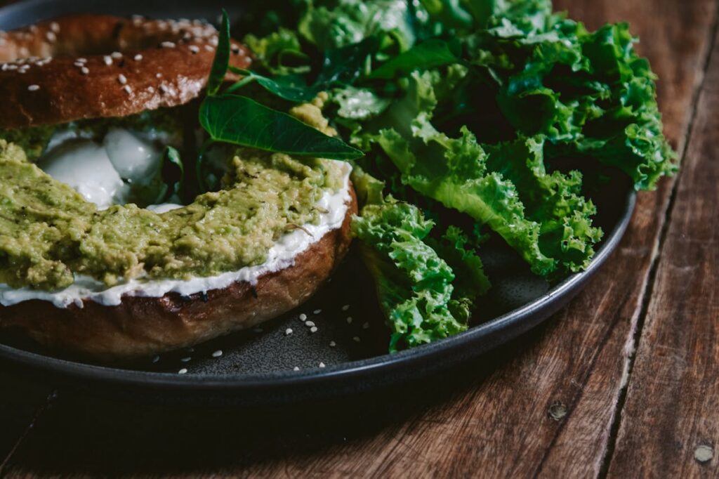 Bagel topped with a green spread and leafy greens on a dark plate placed on a wooden table.