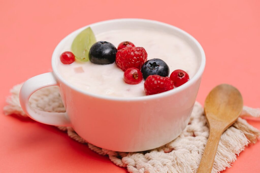 Cup of yogurt can get rid of bloating, topped with raspberries, blueberries, and a lime slice on a pink background with a wooden spoon beside it.
