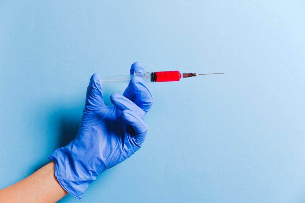 Gloved hand holding a syringe filled with red fluid against a light blue background, test for celiac. How to get rid of bloating.