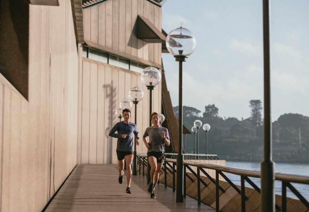 Two people jogging outdoors along a waterfront path beside a building with round lamp posts.