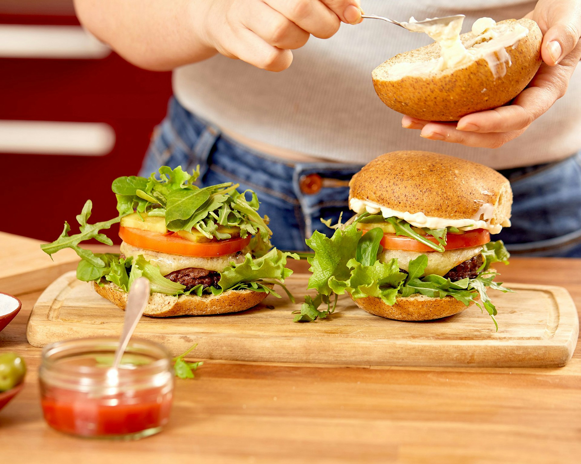 Two assembled burgers with lettuce, tomato, and sauce on a wooden board while a person adds mayonnaise to the top bun of one burger.