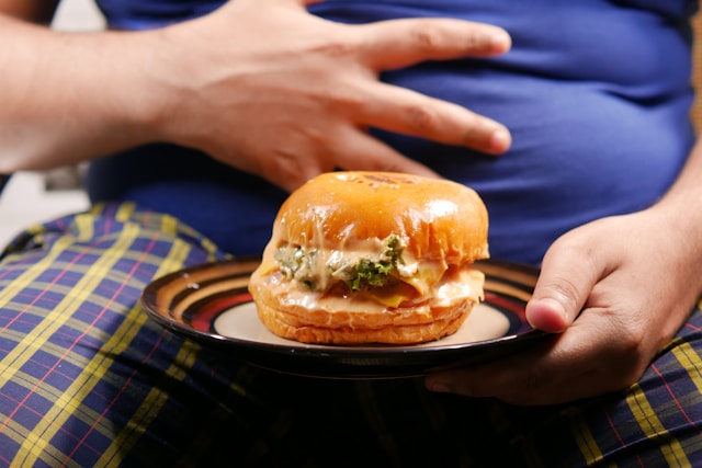 Person holding a plate with a burger while their other hand rests on a visibly bloated stomach. So, how to reduce bloating?