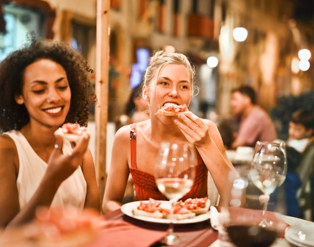 Two women eating at a restaurant table, one taking a bite of food while the other smiles. Swallowing or talking while eating can bring air into the digestive tract.